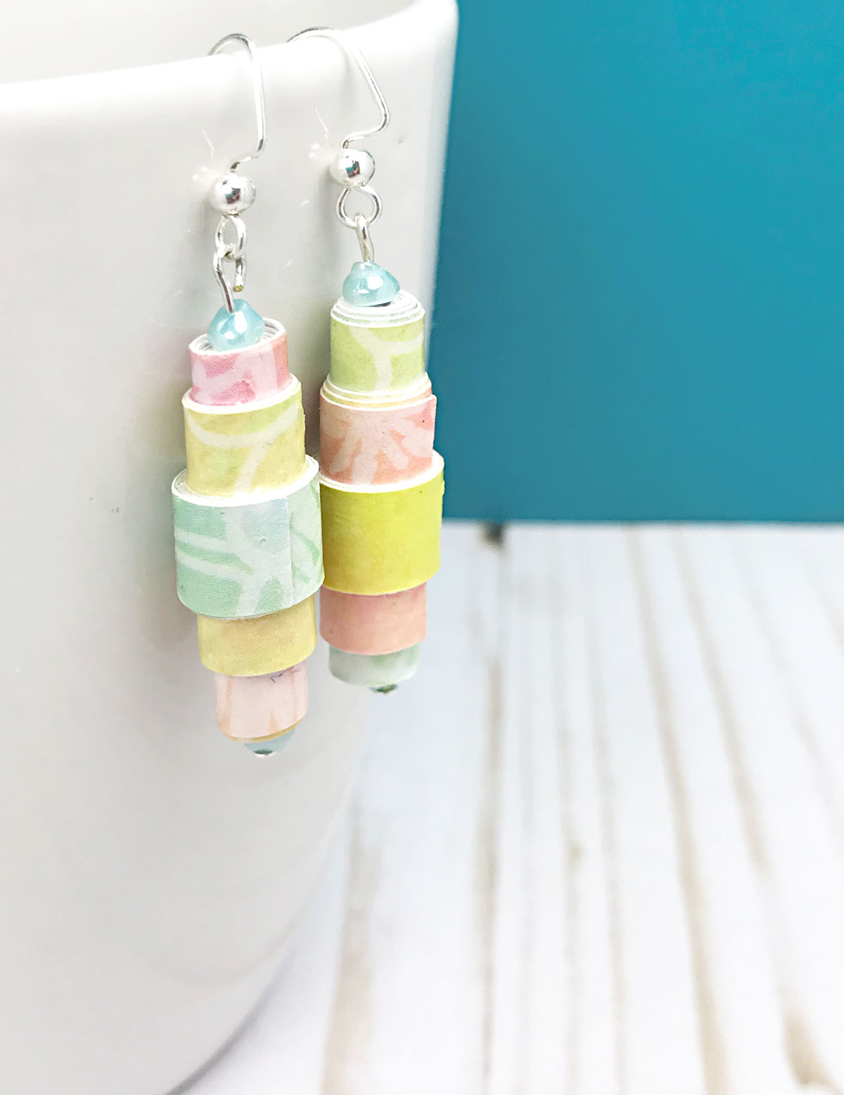 Rainbow paper bead earrings hanging from white cup.